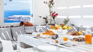A breakfast table with a view of a coastal landscape, including a white table, breakfast items, and a window with a view of the sea and a mountain. - Erossea Villa Santorini Imerovigli (Santorini)
