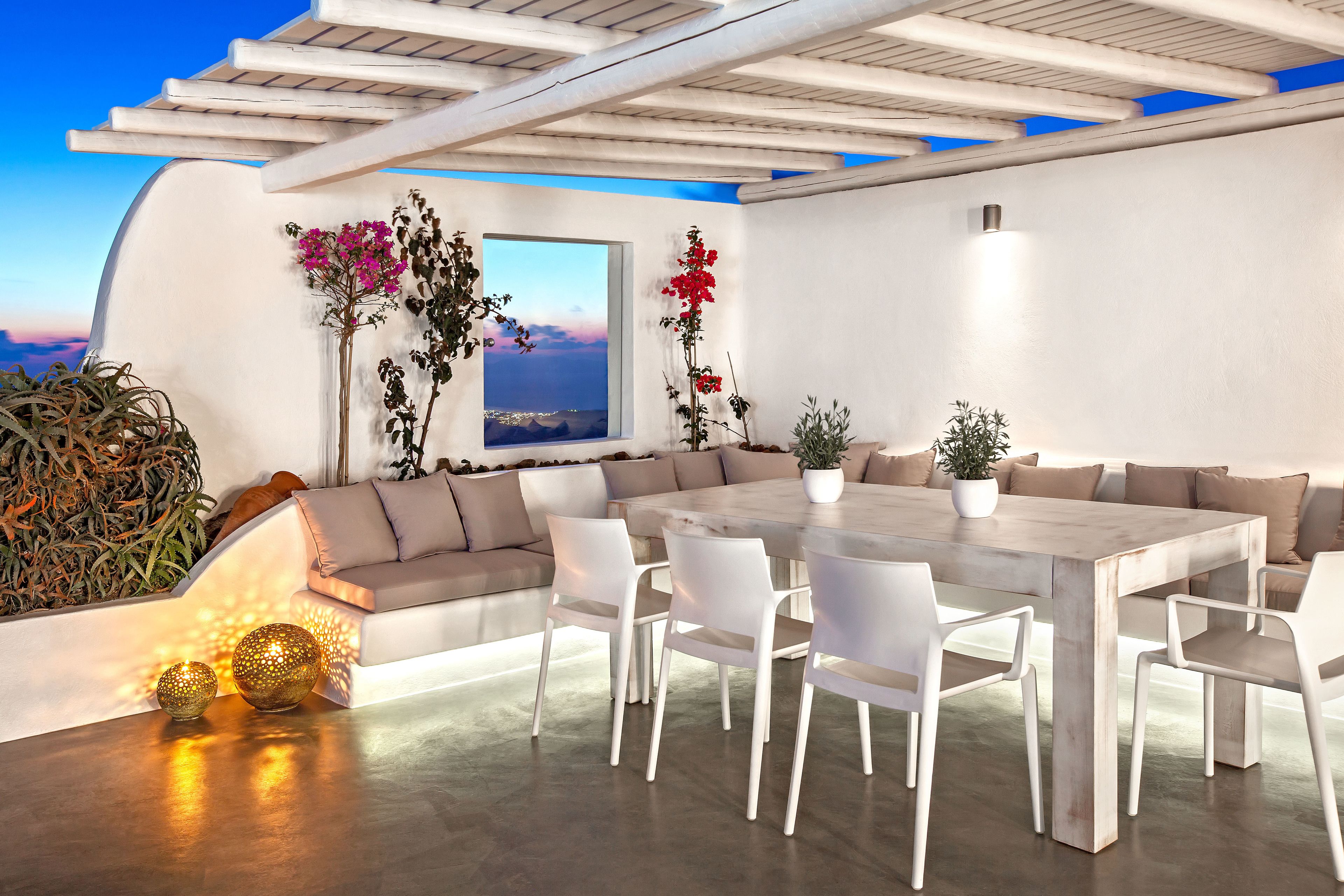 A modern outdoor dining area with a large table and white chairs, surrounded by a white wall and a view of the sea.