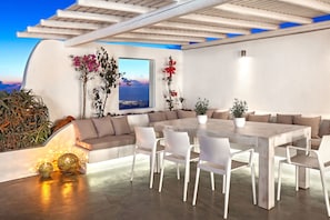 Uma área moderna para refeições ao ar livre, com uma mesa grande e cadeiras brancas, cercada por um muro branco e vista para o mar.