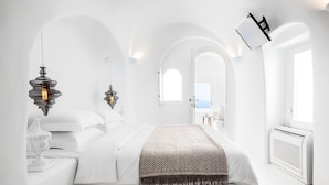 A modern, white-themed bedroom with a bed, two wall-mounted lamps, and a view through an arched doorway. - Erossea Villa Santorini Imerovigli (Santorini)