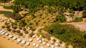 Luchtbeeld van een strandresort met rijen palmbomen, strandparasols en hutten met rieten daken.