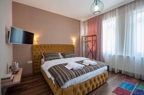 A bedroom with a large bed, a flat-screen TV mounted on the wall, a bedside table with books and a ship model, and a colorful rug. - Newway Apartments (Istanbul)