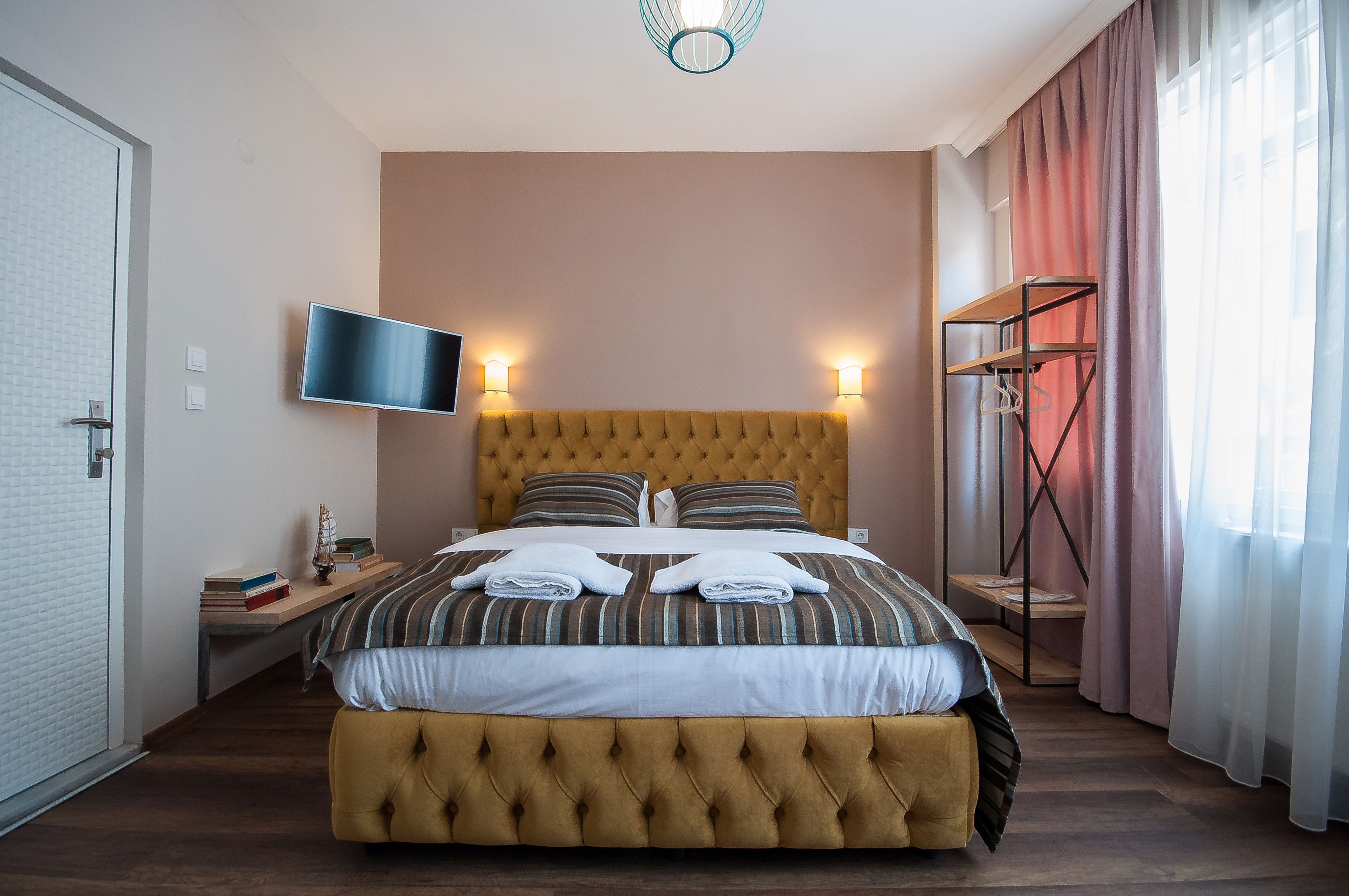 A neatly made bed with a tufted headboard, a bedside table with books and a small decorative item, a wall-mounted TV, and a clothing rack with hanging items.
