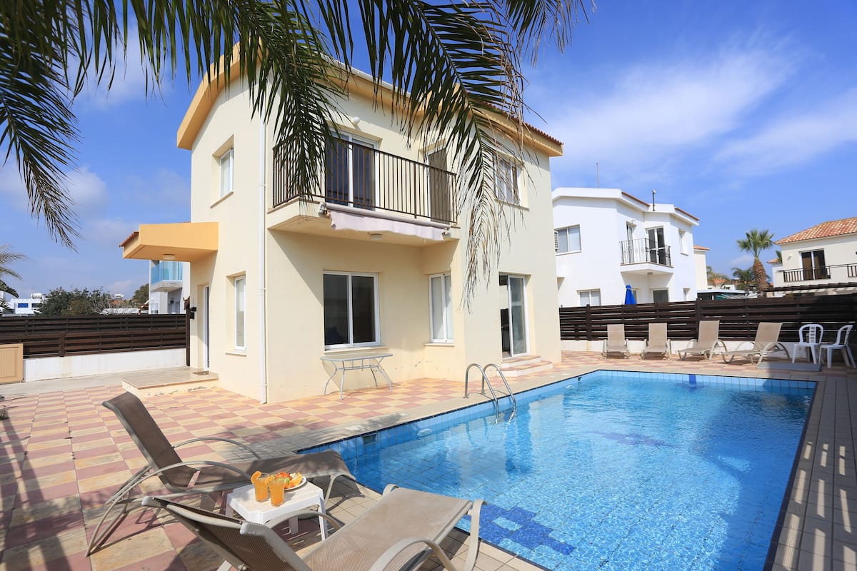 A swimming pool with lounge chairs and a table, surrounded by a building with balconies and a fence.