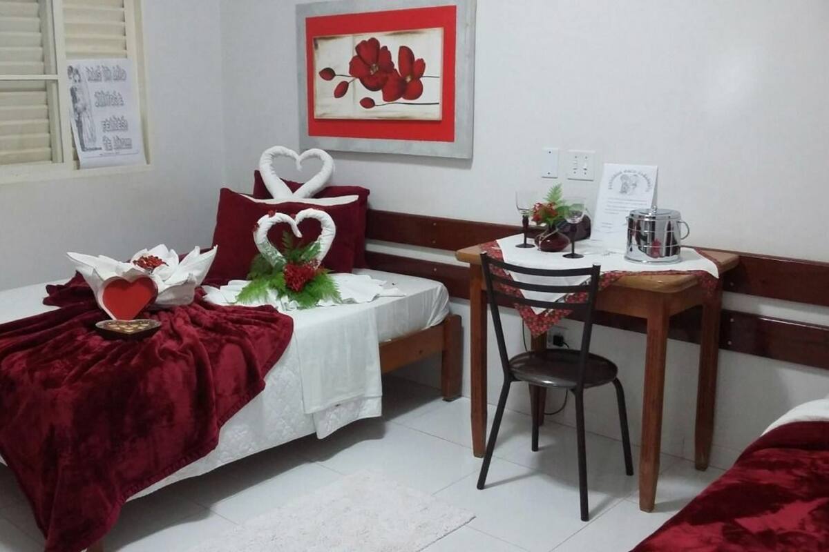 Ein kleiner, ordentlicher Raum mit einem Bett, einem Schreibtisch und einem Stuhl. Auf dem Bett liegt eine herzförmige rote Dekoration und an der Wand hängt ein gerahmtes Bild.