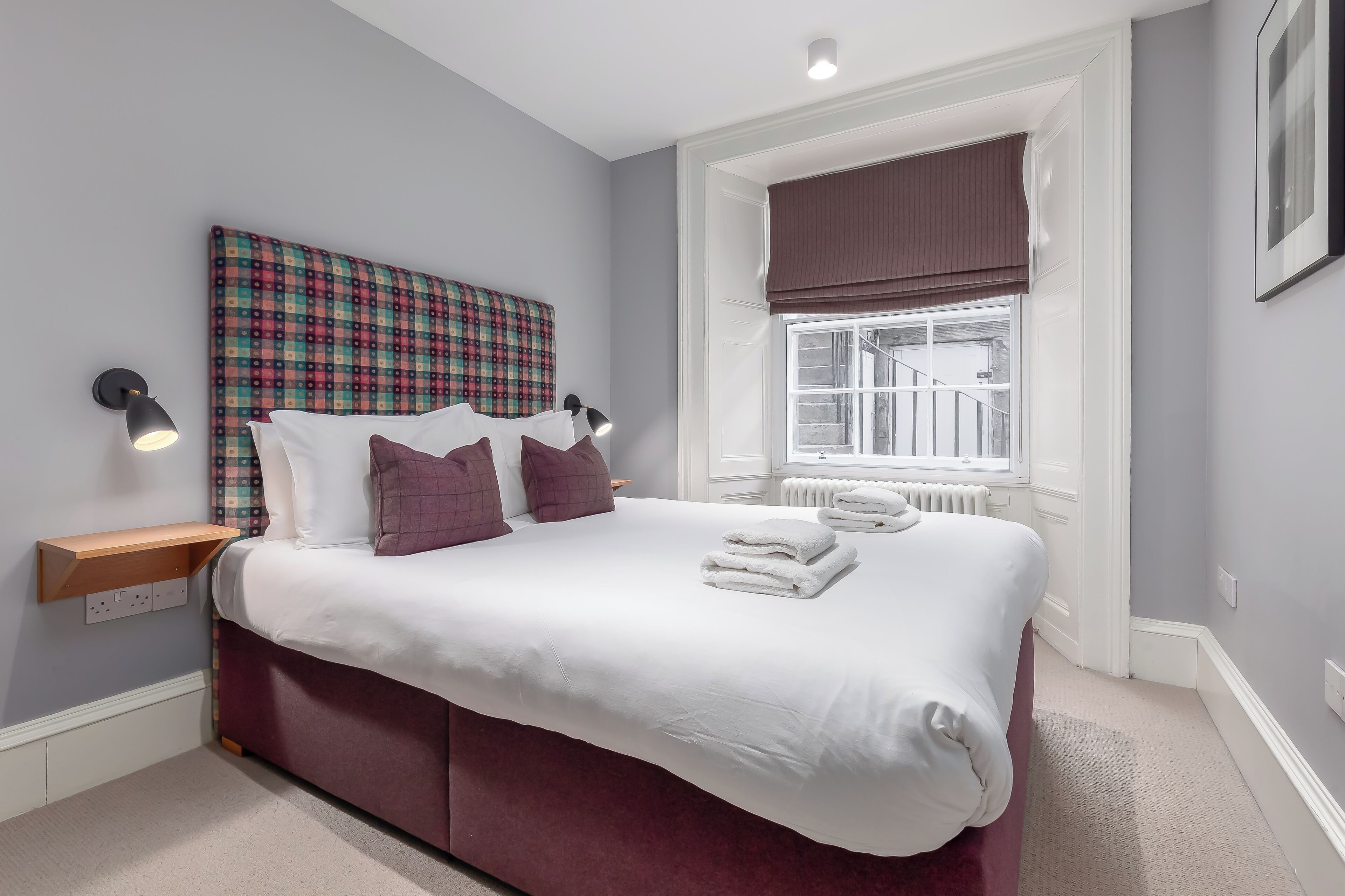 A neatly made bed with white linens and purple pillows, a plaid headboard, and a bedside lamp.