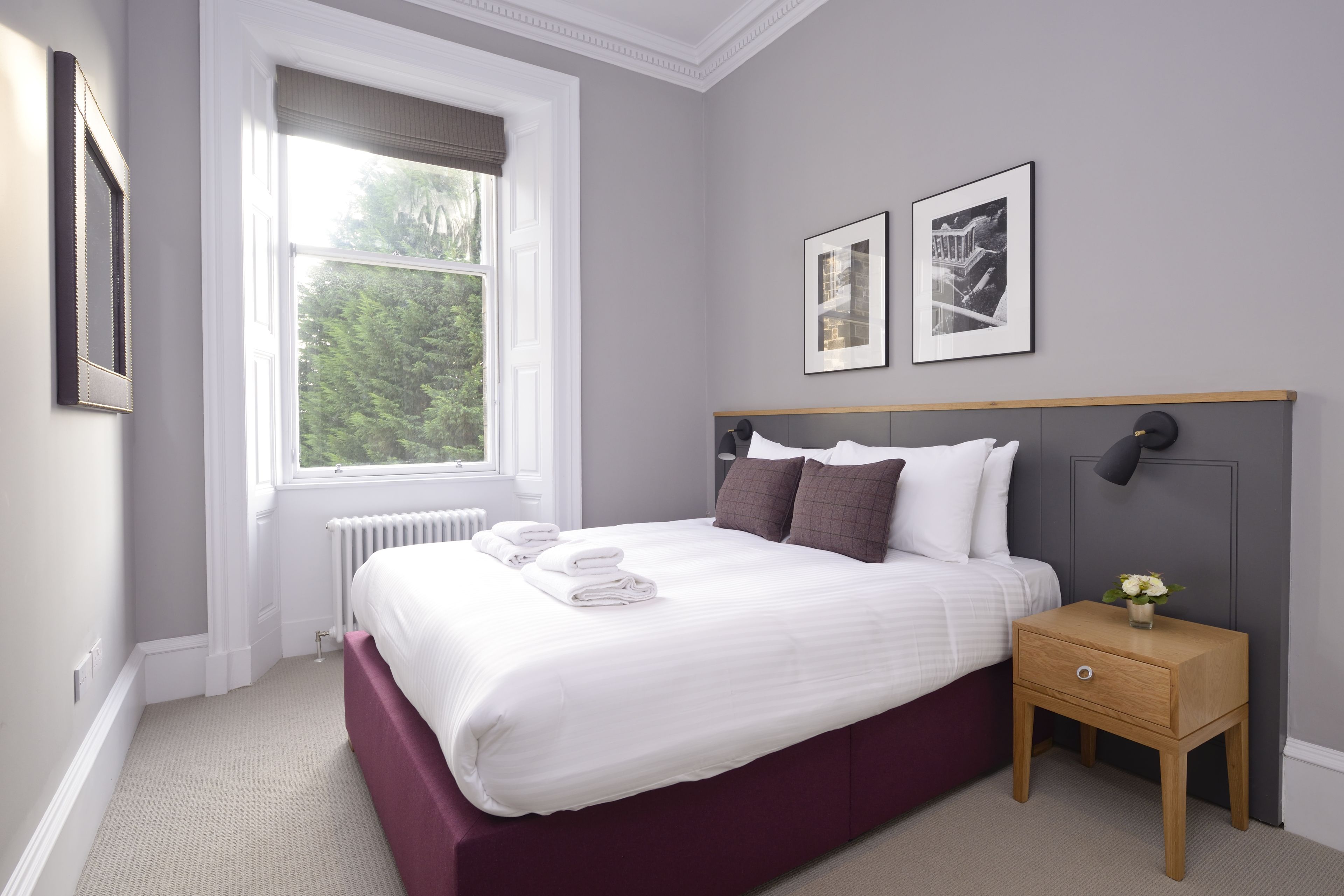 A neatly made bed with white linens and a wooden headboard, a small bedside table with a lamp, and two framed pictures on the wall.