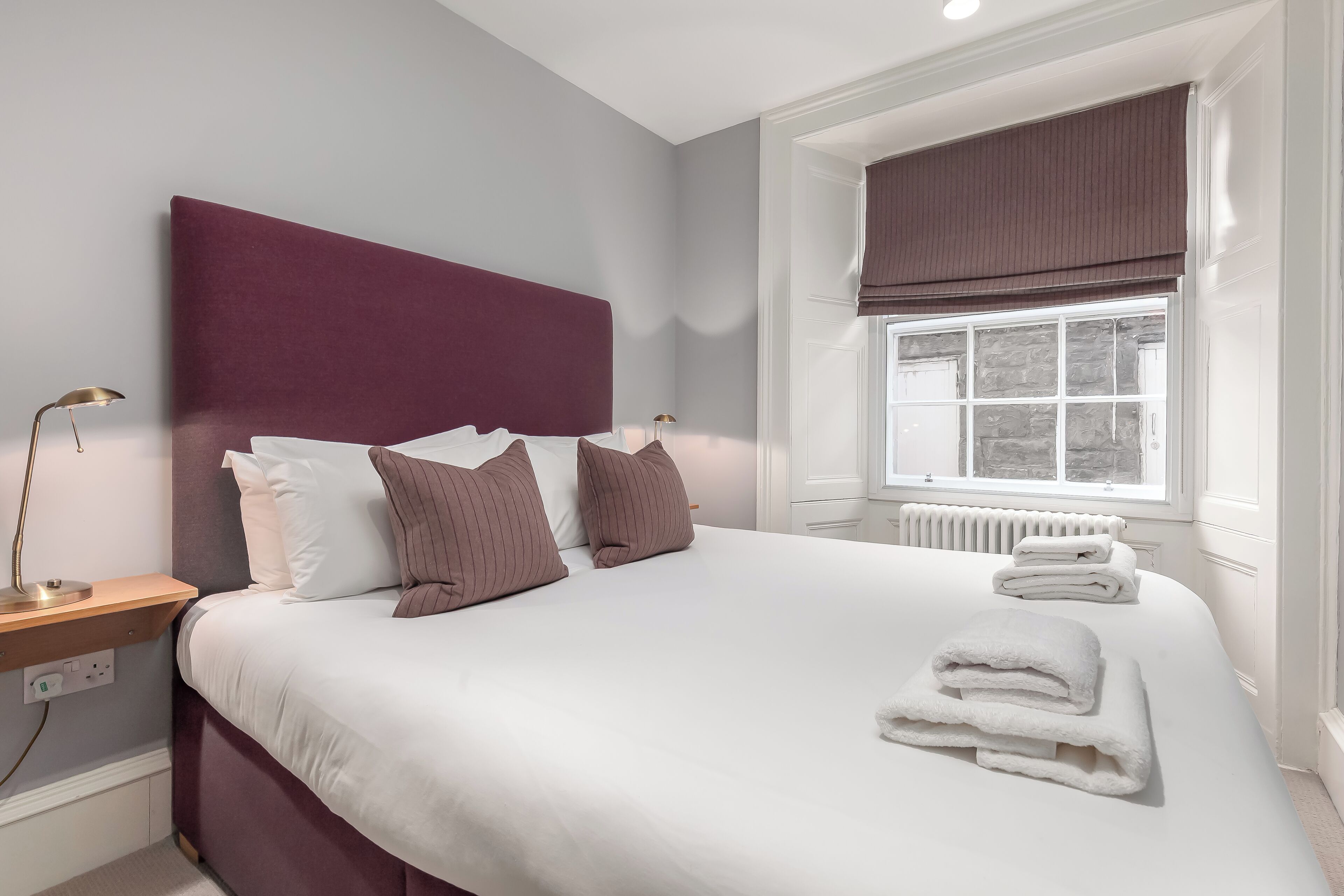 A neatly made bed with white linens and brown pillows, a bedside table with a lamp, and a window with a brown blind.