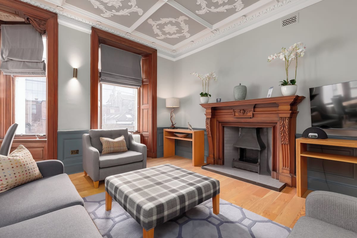 Una sala de estar con sofá, reposapiés, chimenea, televisor y plantas decorativas.
