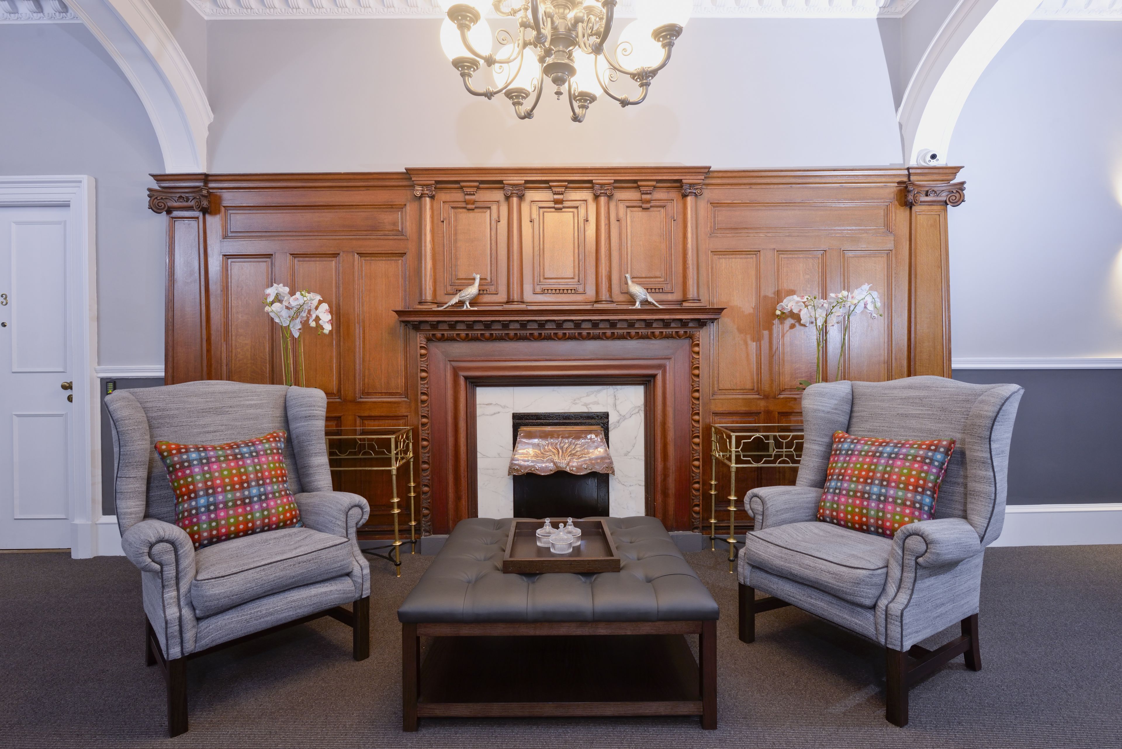 A room with two armchairs, a wooden mantelpiece with a fireplace, and a chandelier.