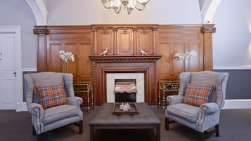 A room with two armchairs, a wooden mantelpiece with a fireplace, and a chandelier.
