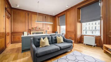 A spacious living room with a grey sofa, wooden paneling, a large window with a view, and a kitchen area in the background.