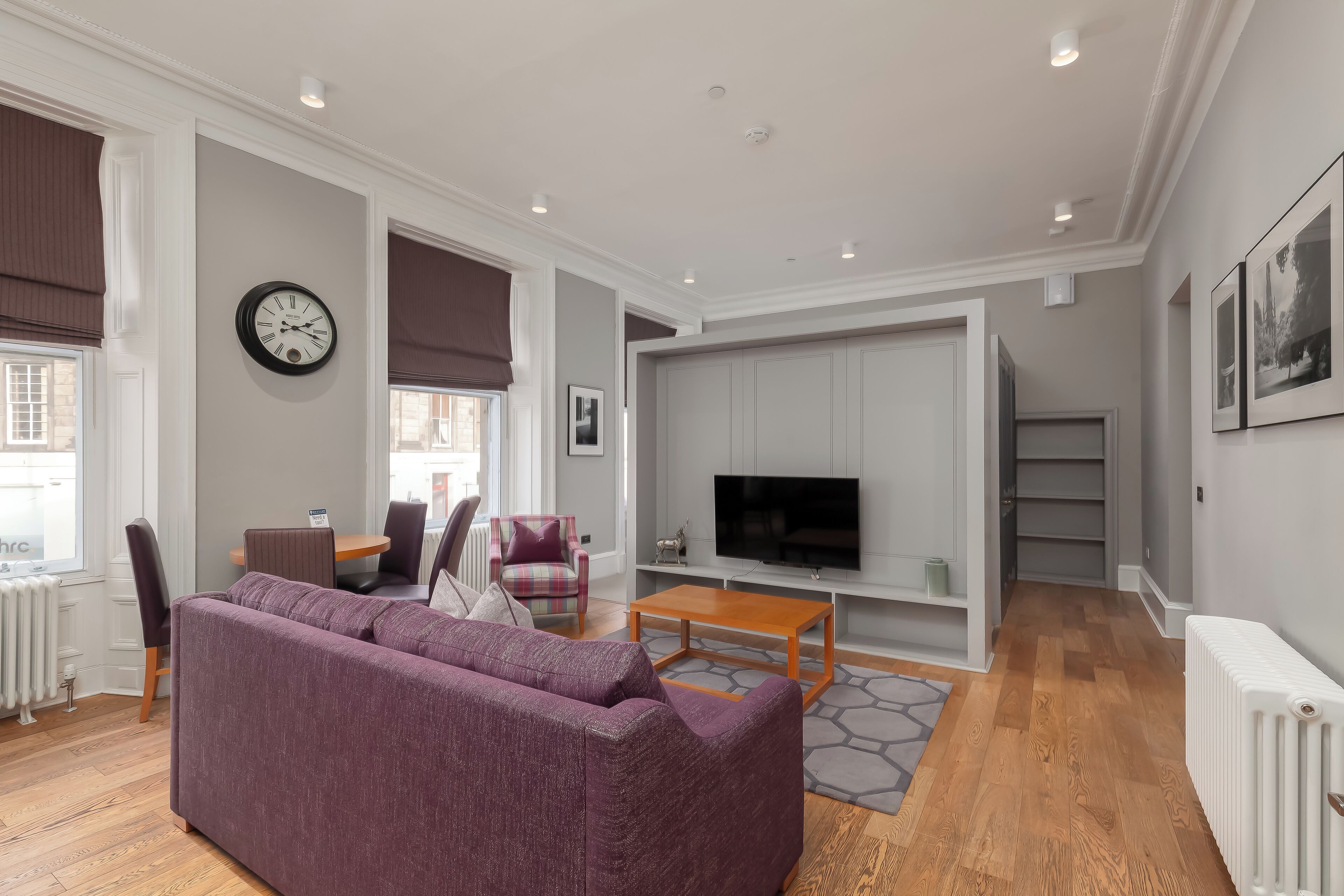 Een moderne woonkamer met een paarse bank, een houten salontafel en een flatscreen televisie aan een grijze muur.