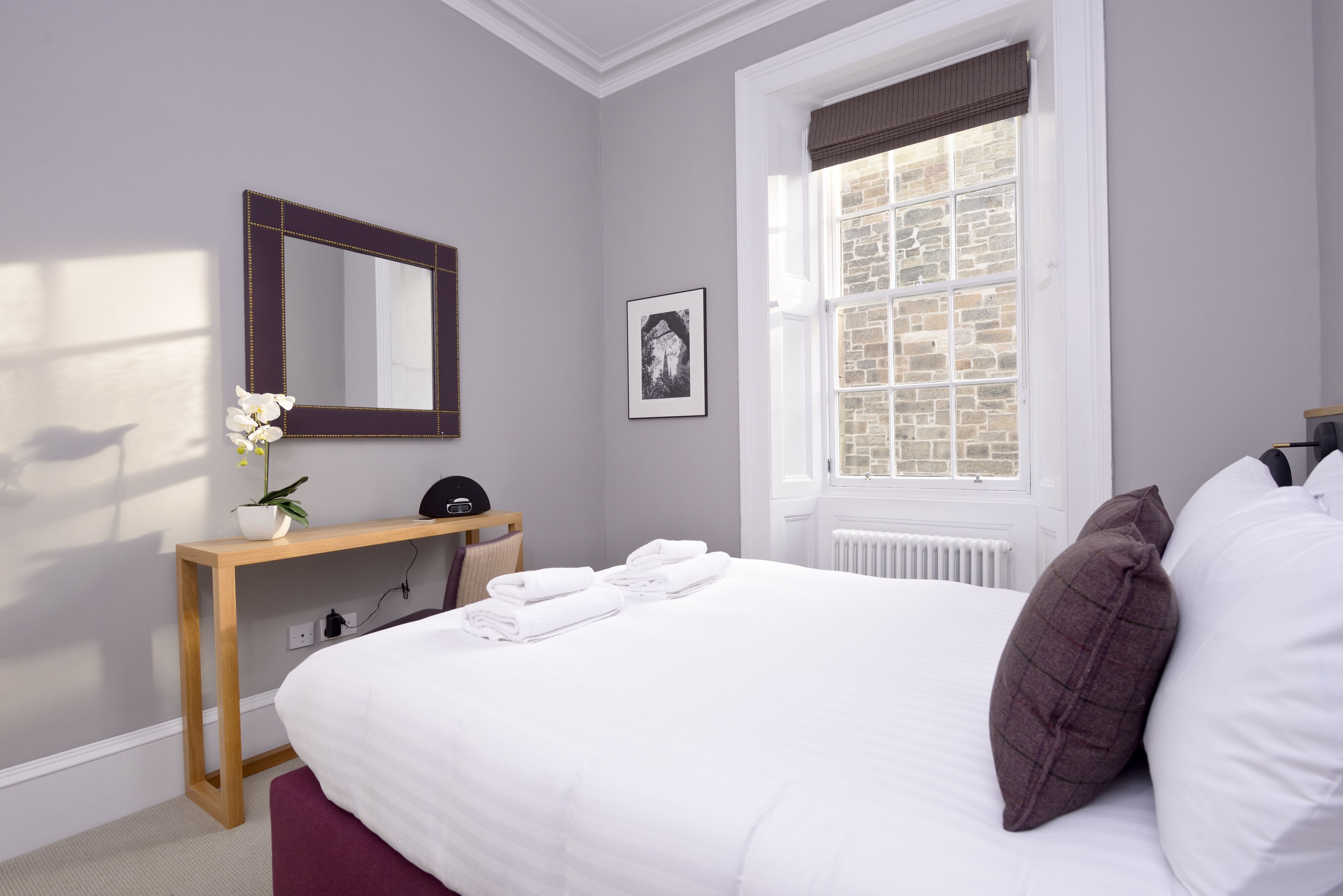 A neatly arranged bedroom with a large bed, a bedside table, a mirror, and a window with a view of a brick wall.
