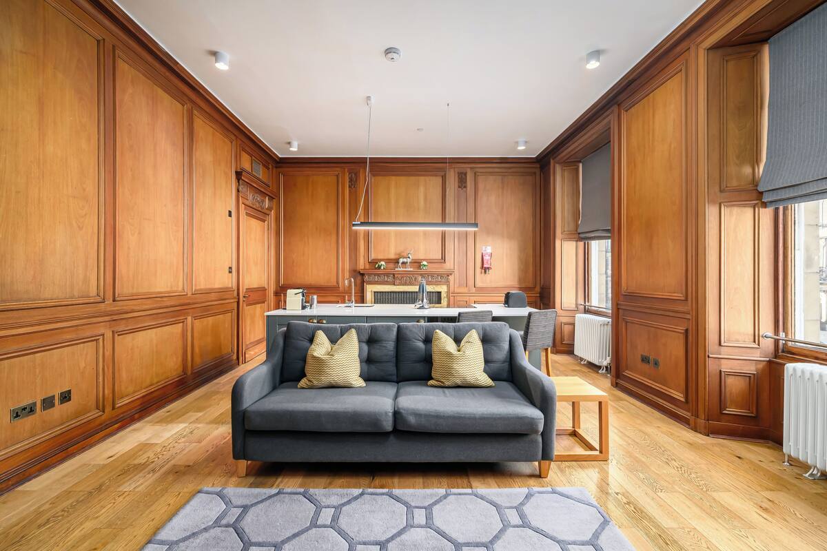 Un salón espacioso con un sofá gris, paneles de madera, una zona de cocina y vistas amplias a través de grandes ventanales.