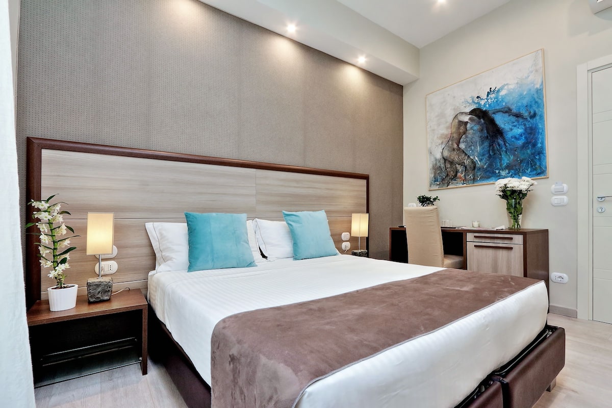 A modern hotel room with a large bed, two bedside tables with lamps, a desk, and a painting on the wall.