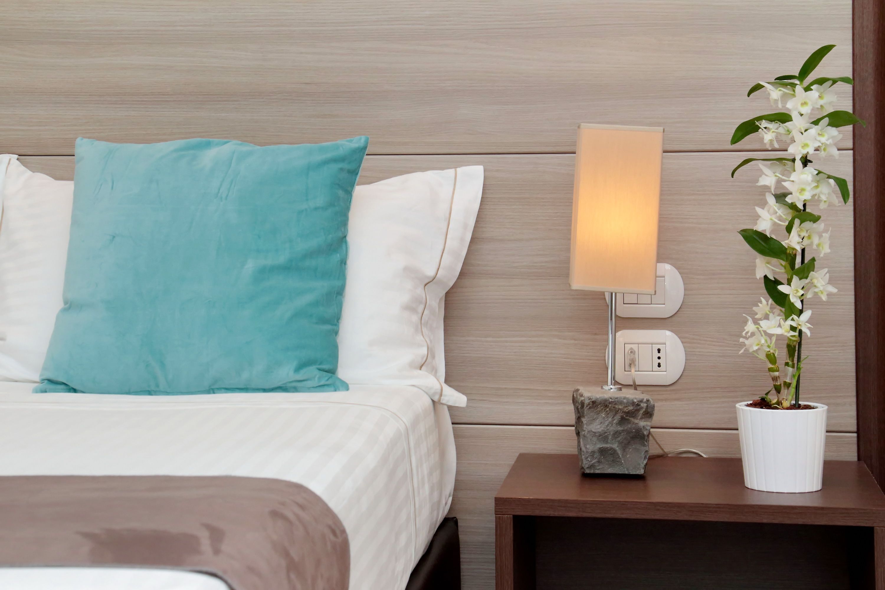 Un letto rifatto con cuscini bianchi e color acquamarina, una lampada da comodino, una piccola pianta in vaso e un comodino con un oggetto simile alla pietra.