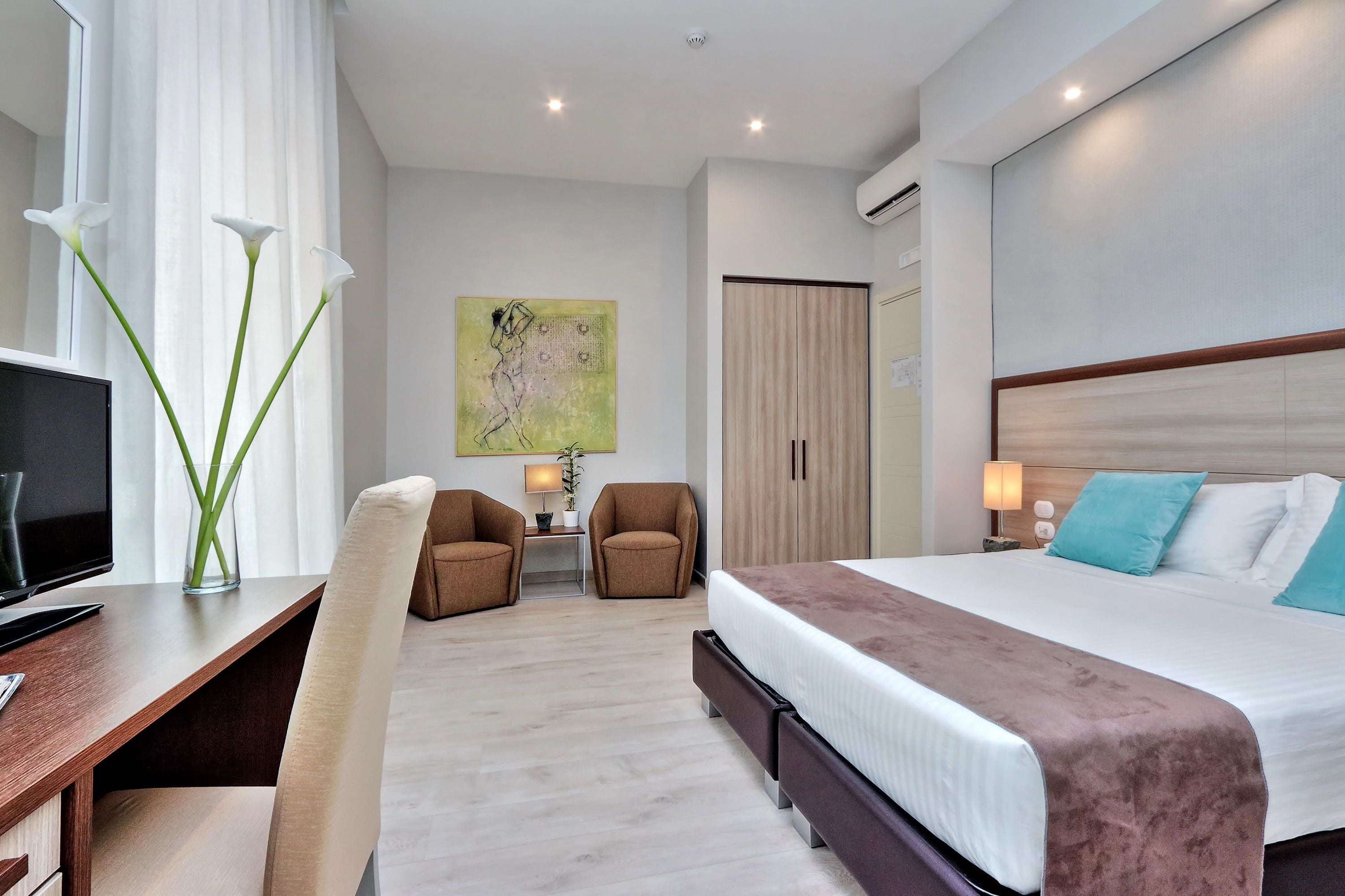 A modern hotel room with a large bed, a desk with a computer, two chairs, a small table with a lamp, and a painting on the wall.