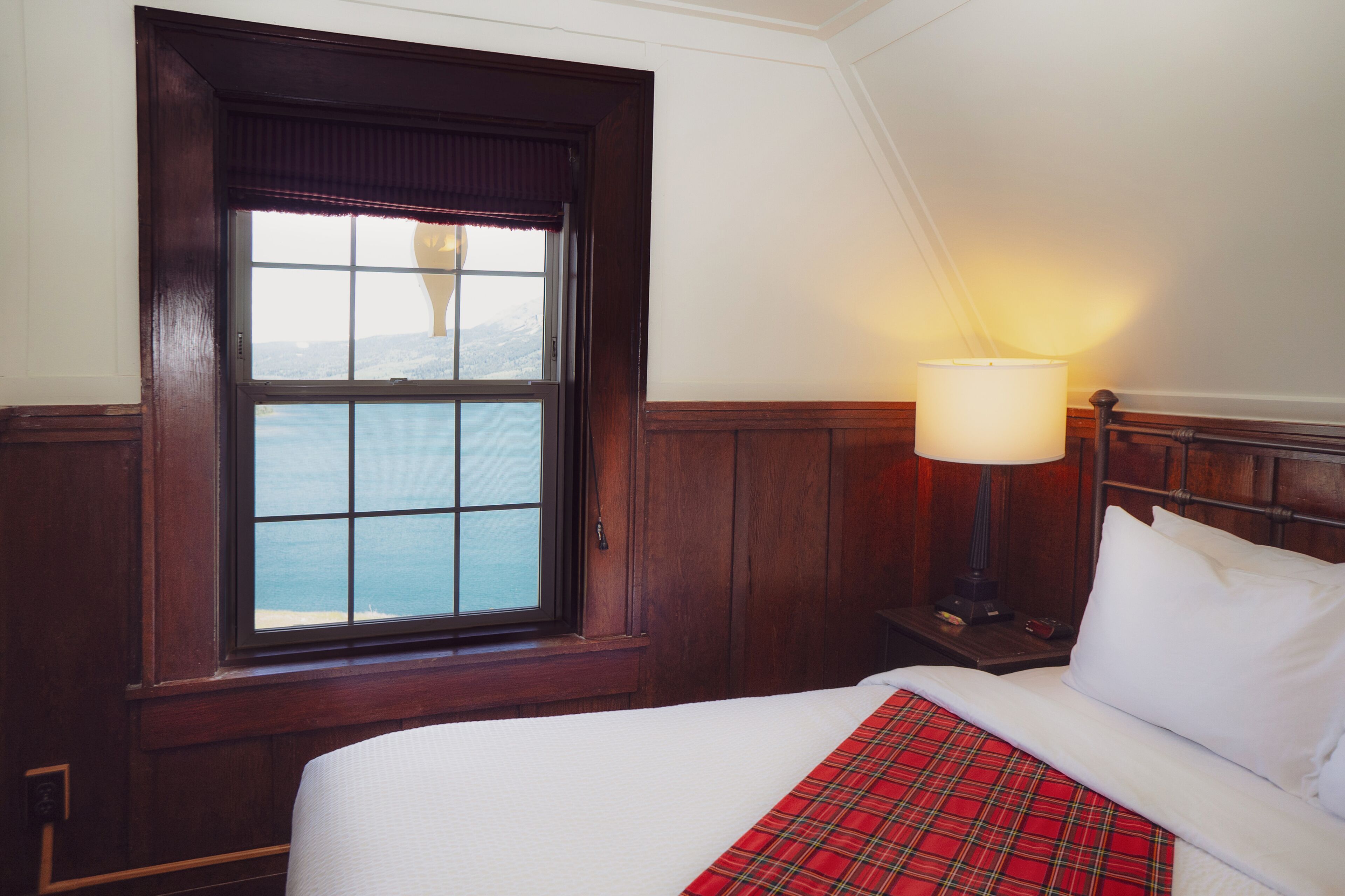 Ein Hotelzimmer mit einem großen Fenster, einem Bett mit weißer Bettwäsche, einer roten Karo-Decke und einem Nachttisch mit einer Lampe.