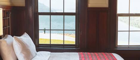 Uma cama com roupa de cama branca e um cobertor xadrez vermelho, uma janela com vista para a praia e montanhas, e painéis de madeira nas paredes.