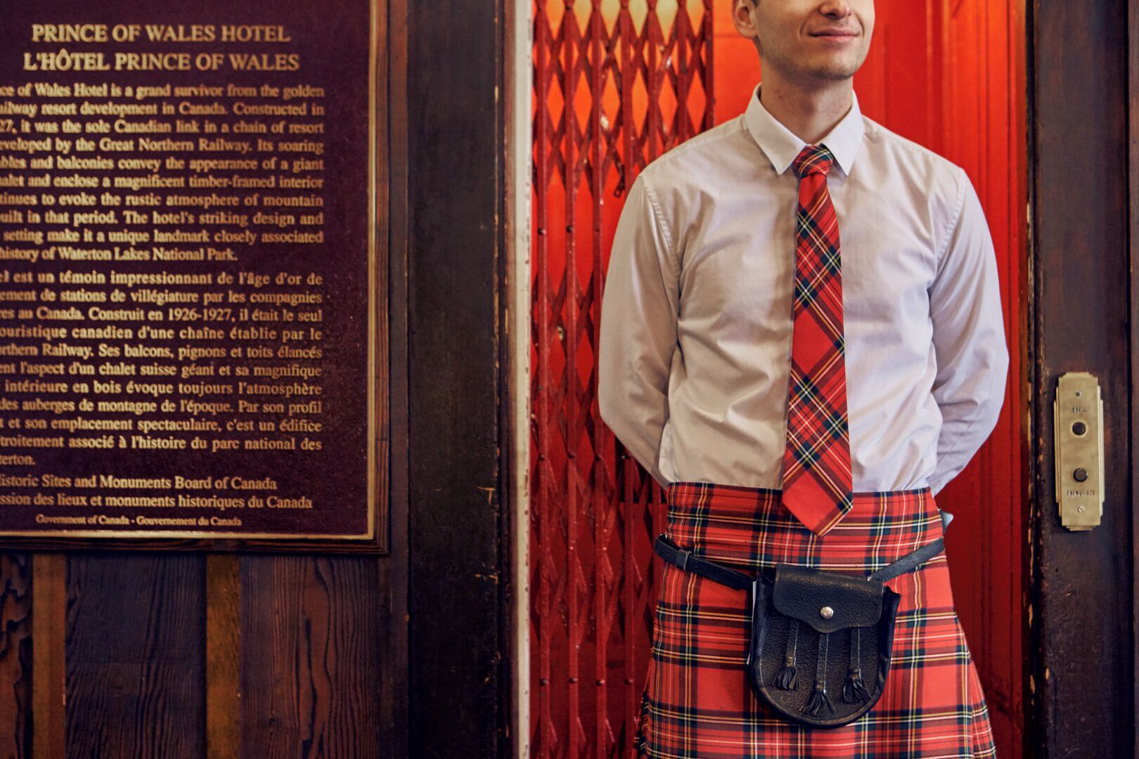 Un homme vêtu d’une chemise blanche et d’un kilt à carreaux rouges se tient devant une porte rouge sur laquelle est affiché un panneau indiquant l’hôtel Prince of Wales.