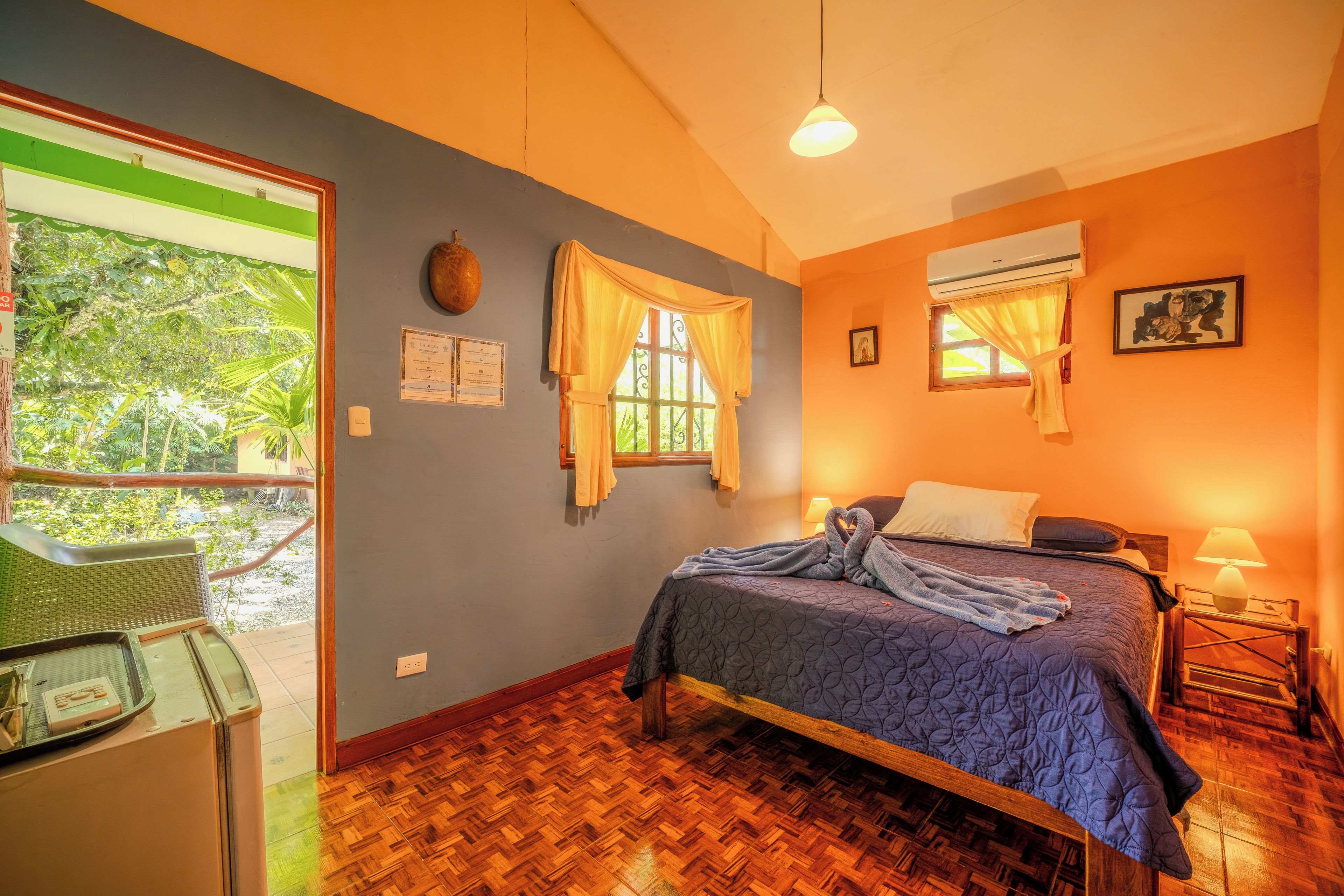 A room with a bed, a window with curtains, a wall-mounted air conditioner, and a view of greenery outside.