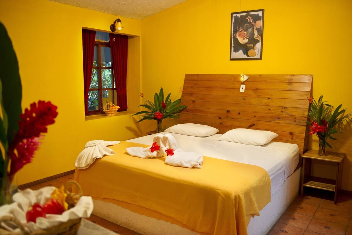 A hotel room with a wooden headboard, a bed with white linens, a yellow bedspread, two bedside tables with lamps, and a window with red curtains.
