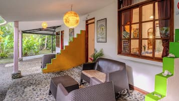 A room with a staircase, wicker furniture, a hanging light fixture, and a large window with curtains.