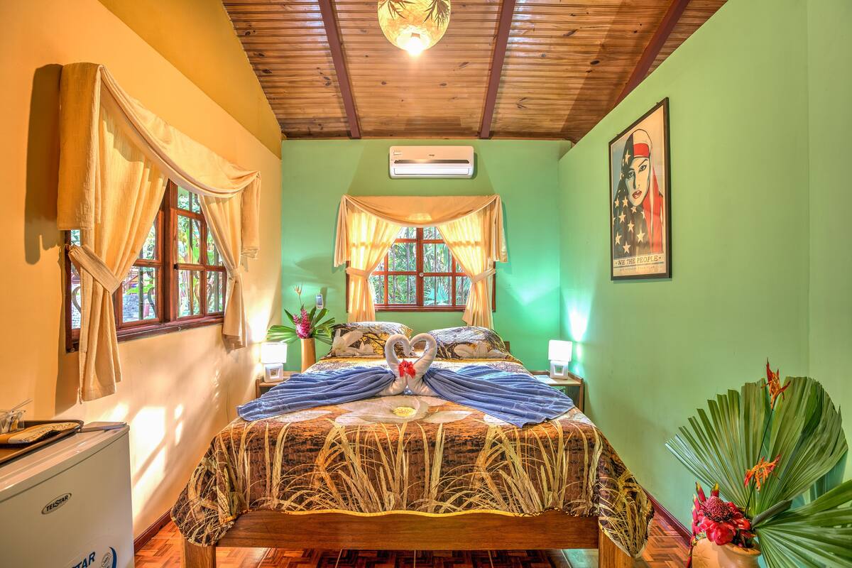 A room with a bed, a ceiling fan, a window with curtains, a wall-mounted air conditioner, and a poster on the wall.