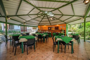 An outdoor dining area with tables and chairs, a kitchen in the background, and a tiled floor.