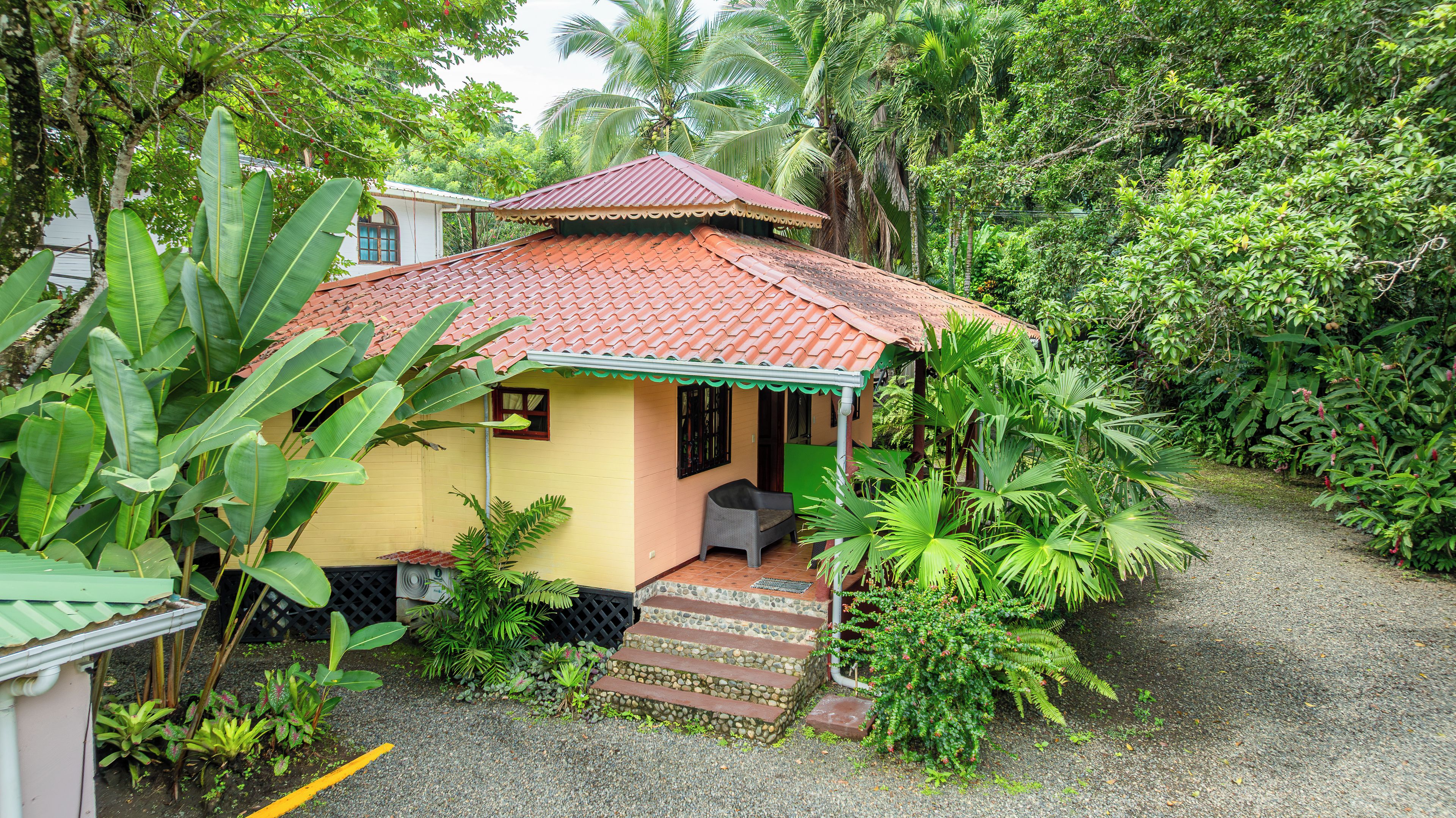 A small house with a tiled roof, surrounded by lush greenery and a gravel path leading to the entrance.