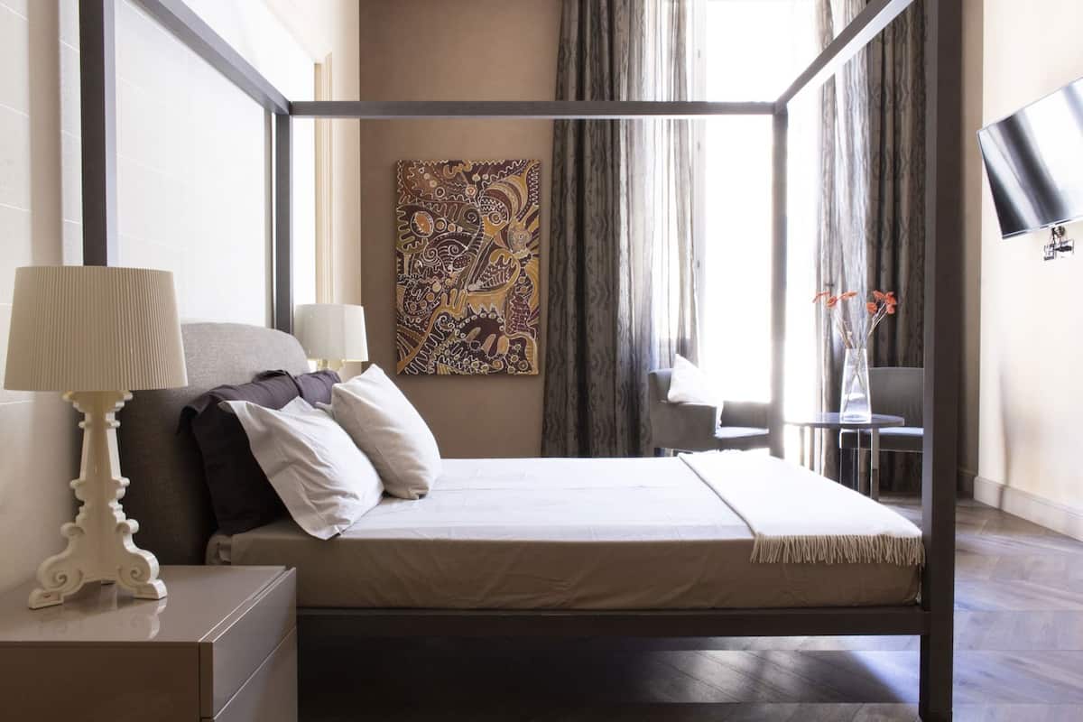 A four-poster bed with a bedside table, a lamp, a chair, a desk, and a painting on the wall.