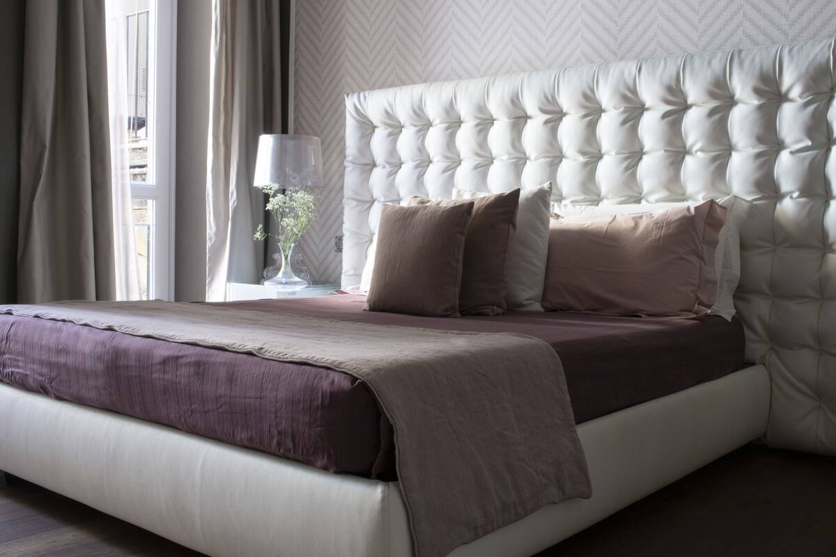 A neatly made bed with a tufted headboard, a bedside table with a lamp, and a window with curtains.