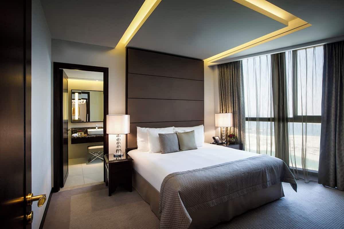Une chambre d’hôtel avec un grand lit, des lampes de chevet, une table de nuit, un miroir et une vue sur l’extérieur grâce à de grandes fenêtres.