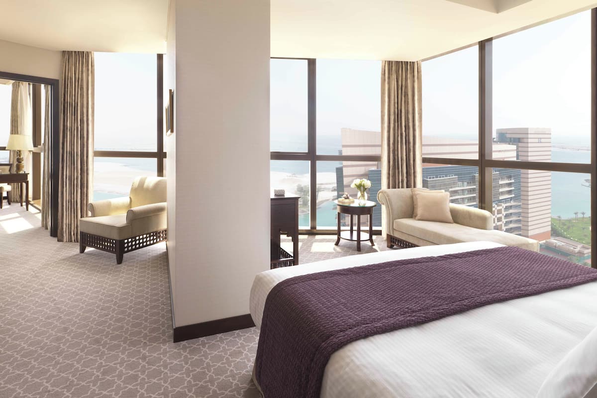 Une chambre d’hôtel avec un grand lit, deux canapés, une petite table agrémentée de fleurs et une vue sur la ville et l’eau.