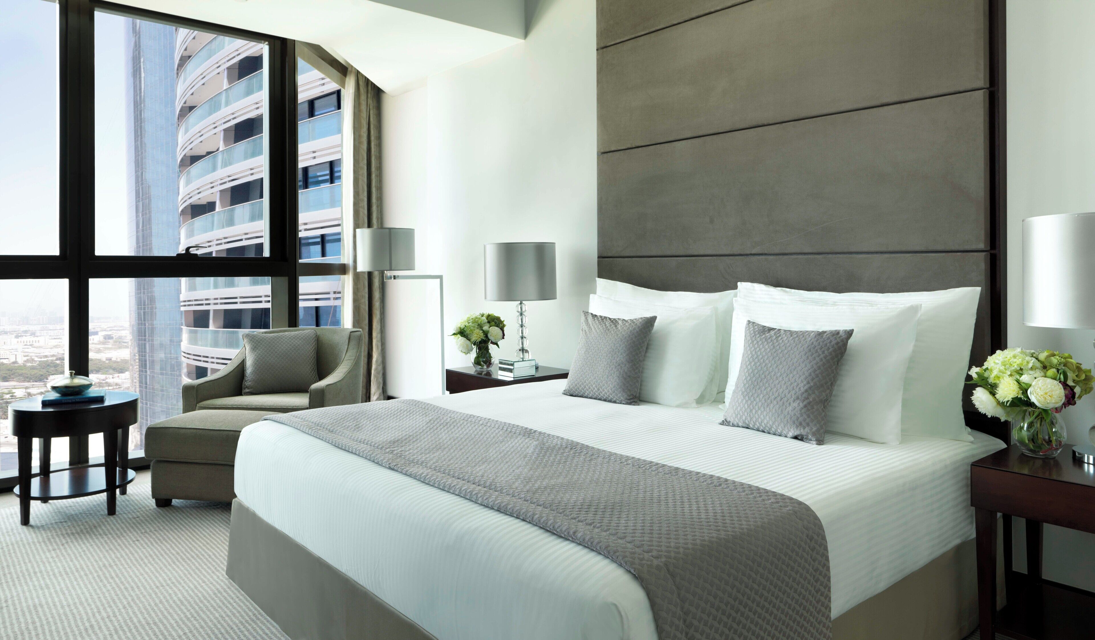 Habitación de hotel moderna con una cama grande, zona de estar, mesita de noche y vistas a un paisaje urbano a través de amplios ventanales.
