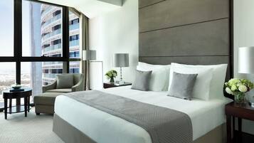 Habitación de hotel moderna con una cama grande, zona de estar, mesita de noche y vistas a un paisaje urbano a través de amplios ventanales.