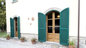 Un bâtiment doté de deux fenêtres en arc surmontées de volets verts, d’une façade en pierre et de plantes en pot à l’entrée.