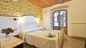 Une chambre avec un mur en pierre, un plafond en bois, un lit recouvert d’une courtepointe, une table de chevet ornée d’un vase et une fenêtre offrant une vue agréable.
