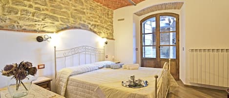 Un dormitorio con pared de piedra, techo de madera, cama con colcha, mesita de noche con un jarrón y ventana con vistas.