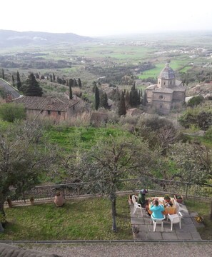 A terrace with a table and chairs overlooking a rural landscape with a church and houses. - Appartamento Luna (Cortona)