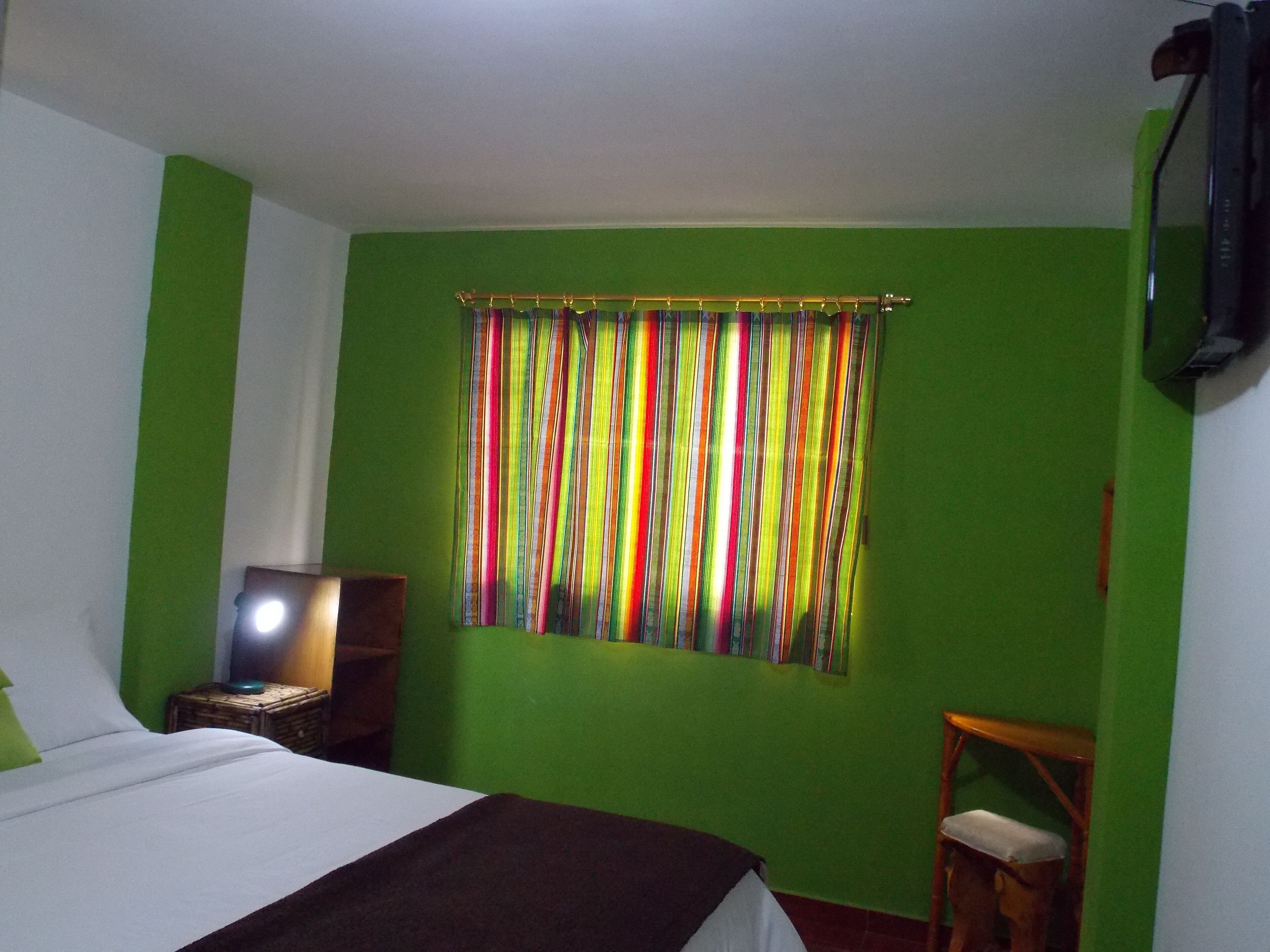 Una habitación con cama, una mesita de noche con lámpara, una silla y un ventanal con cortinas de colores.