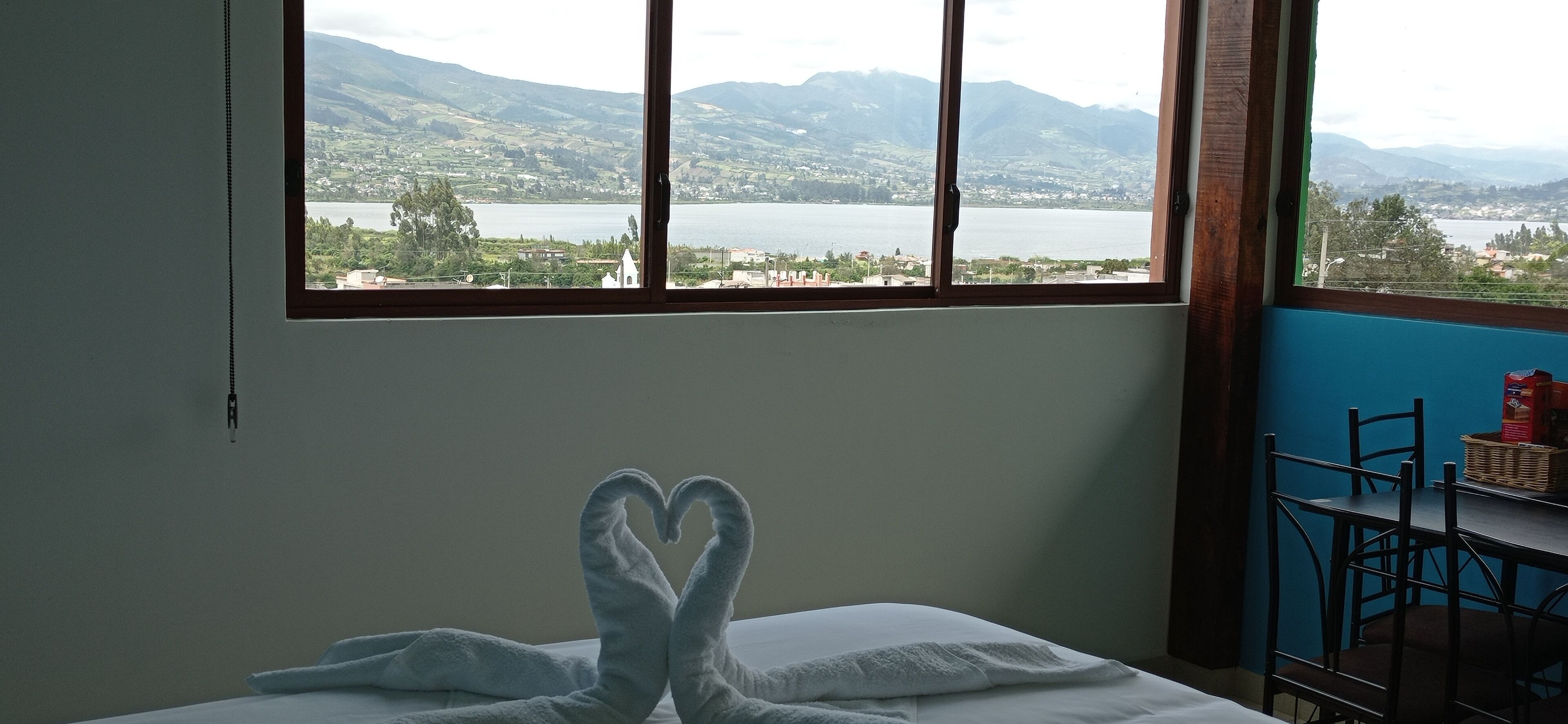 A room with a bed, a window with a view of a lake and mountains, and a table with chairs.