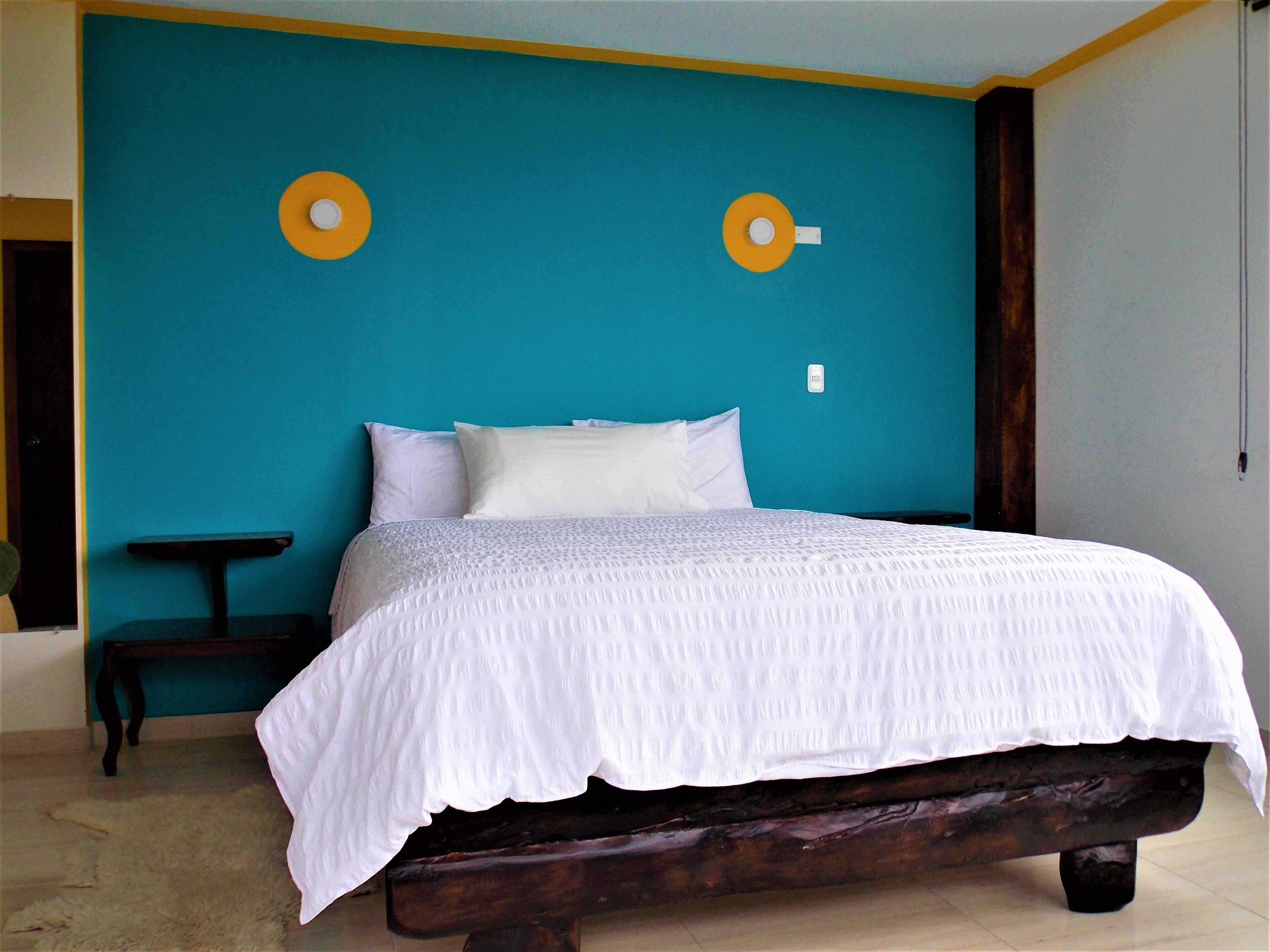 Una cama con cubrecama blanco en una habitación con paredes color turquesa y un banco de madera.