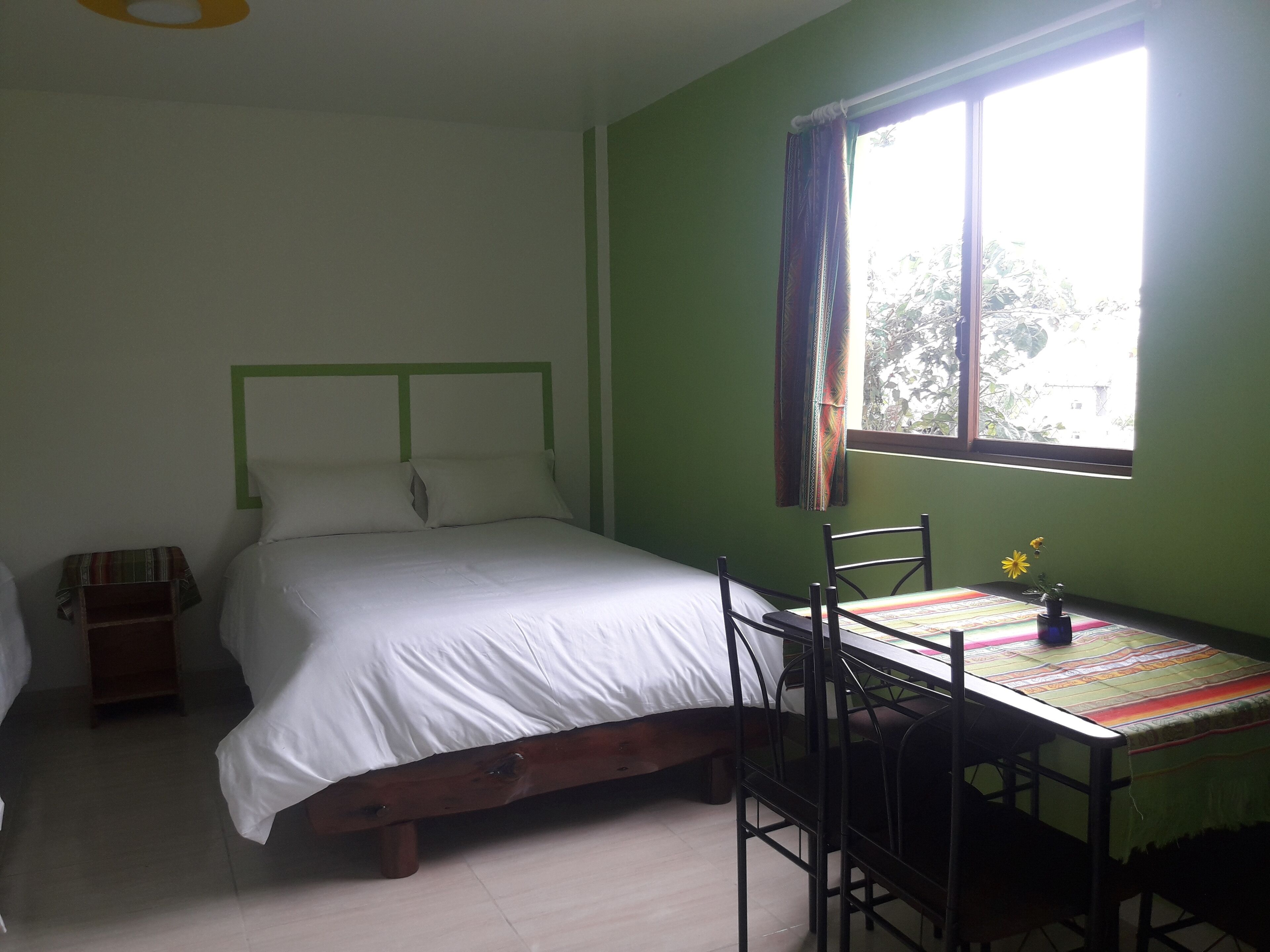A bedroom with a bed, a table with chairs, a window with curtains, and a small table with a vase of flowers.