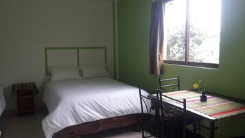 A bedroom with a bed, a table with chairs, a window with curtains, and a small table with a vase of flowers.