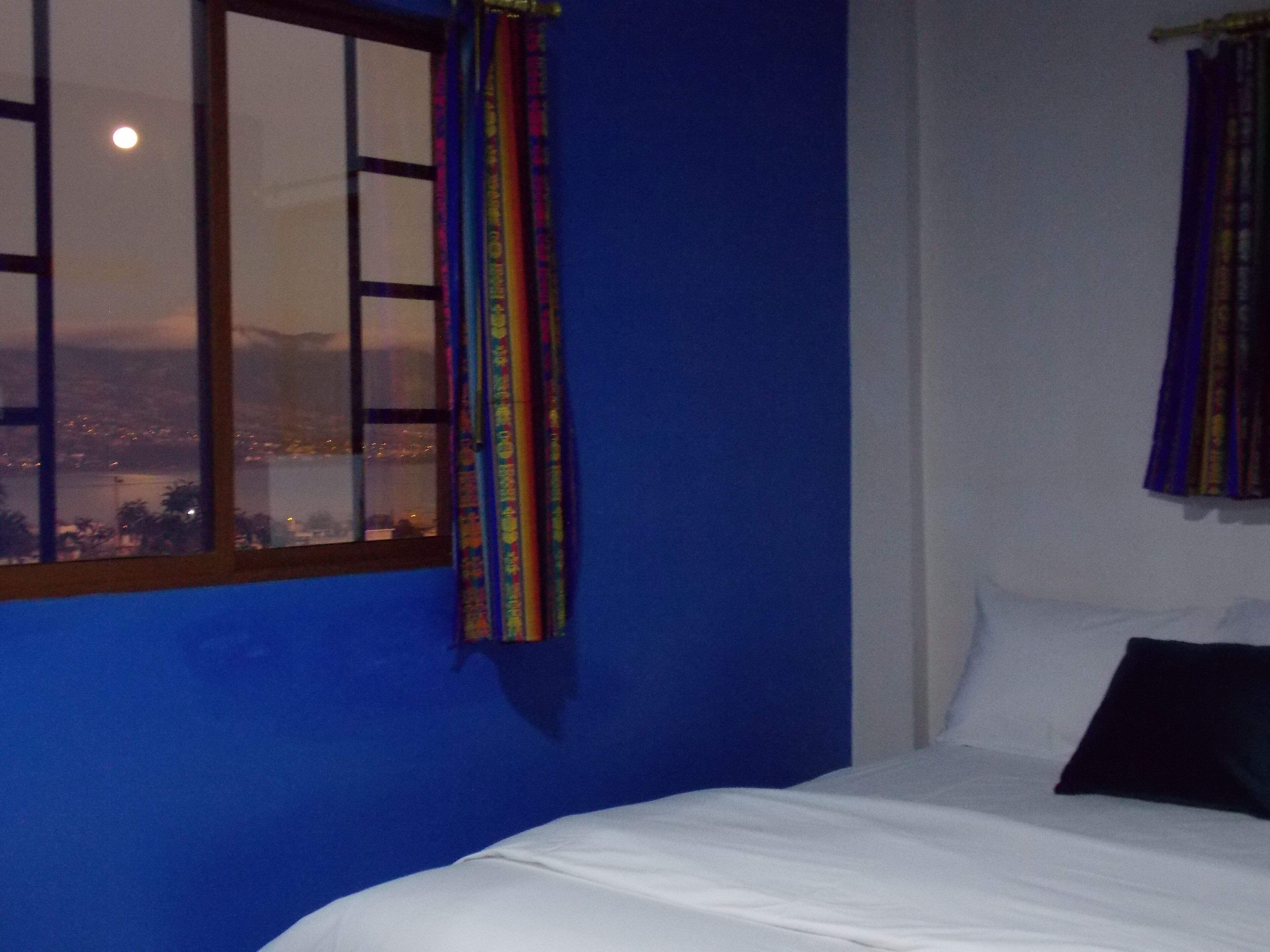Een kamer met een bed, een raam met gordijnen en uitzicht op een stadsbeeld en bergen.