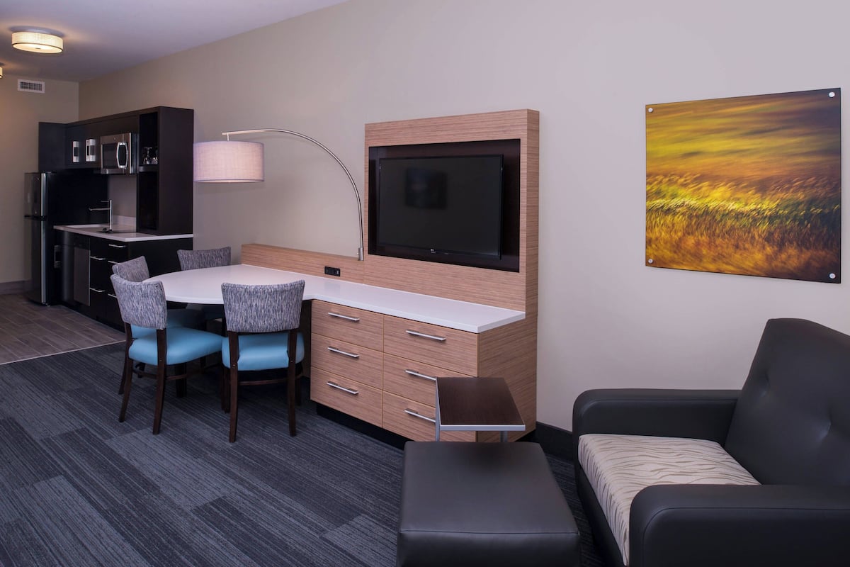 A compact hotel room with a kitchenette, a dining area with a table and chairs, a flat-screen TV mounted on the wall, and a large window.