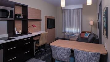 A modern hotel room with a kitchenette, a dining area with a table and chairs, a sofa, a TV, and a lamp.