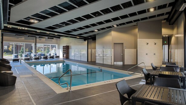 An indoor swimming pool with clear water, surrounded by lounge chairs and tables, and a view of the outside through large windows. - TownePlace Suites by Marriott Saskatoon (Saskatoon)